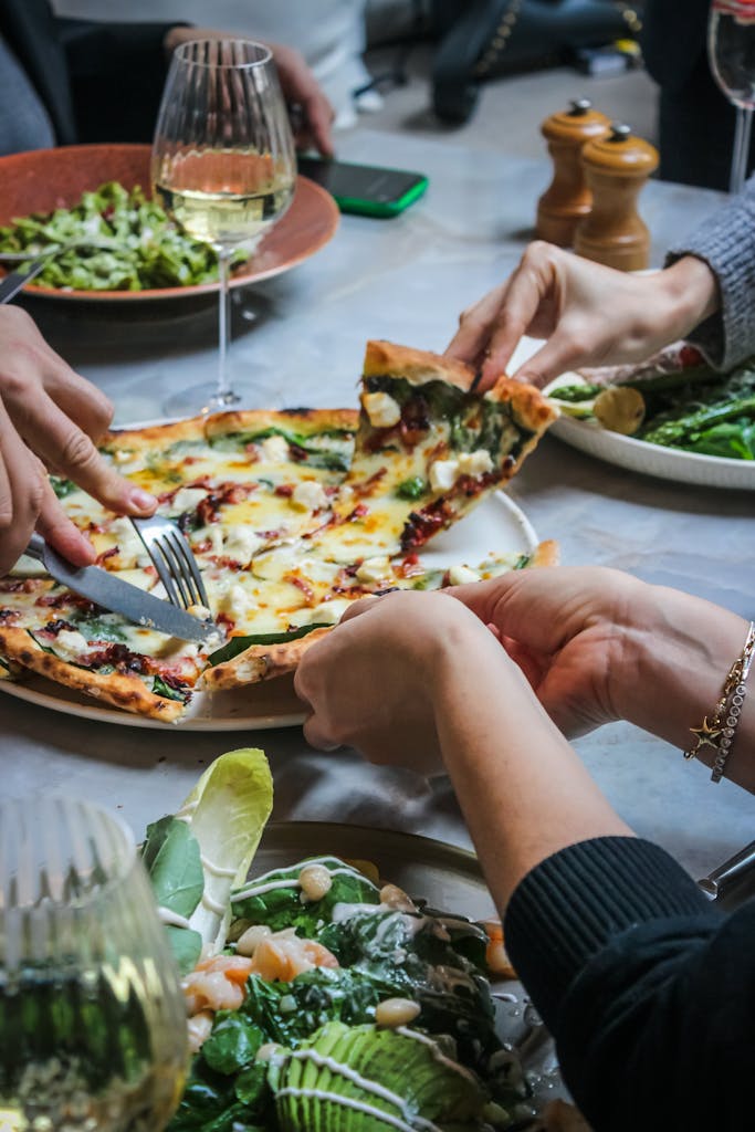 Close-up of hands sharing pizza with salad and wine at a dining table, creating a warm social atmosphere.